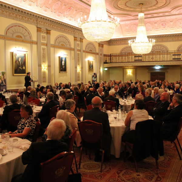 A dinner at Plaisterers' Hall. Thirty circular tables, with white table cloth, beneath a ceiling of chandeliers.