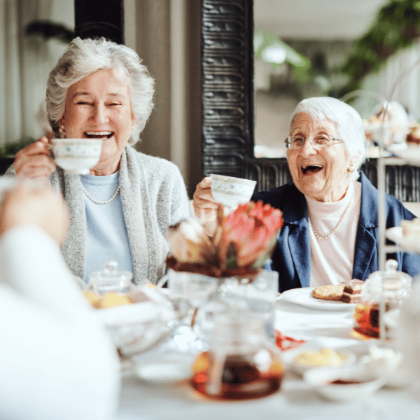 Two elderly women, raising a cup of tea and laughing.