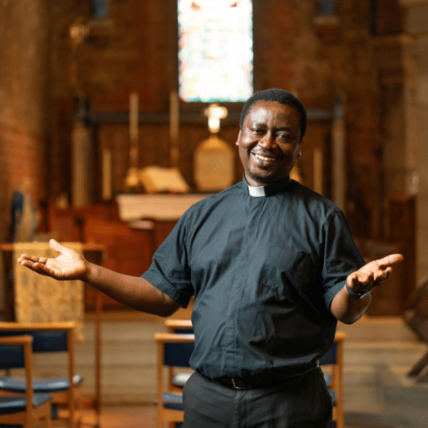 Josiah is wearing a black shirt and a white clerical collar. He is standing in his church, in front of a stained glass window, with his hands outstretched.