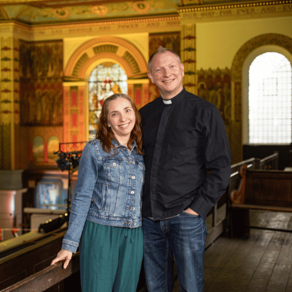 Sara and Graham, stood inside a church. Stained glass windows can be seen behind them.