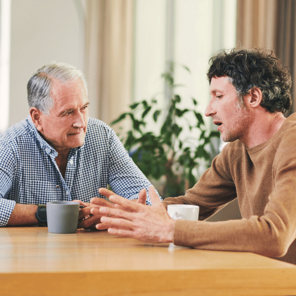 Two men, sat at a table with a hot drink, in conversation.