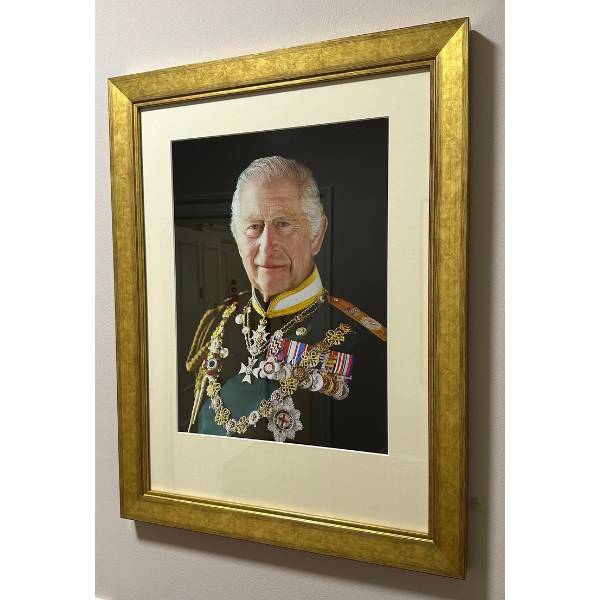 A portrait of King Charles III, in a golden frame, hanging on a white wall.