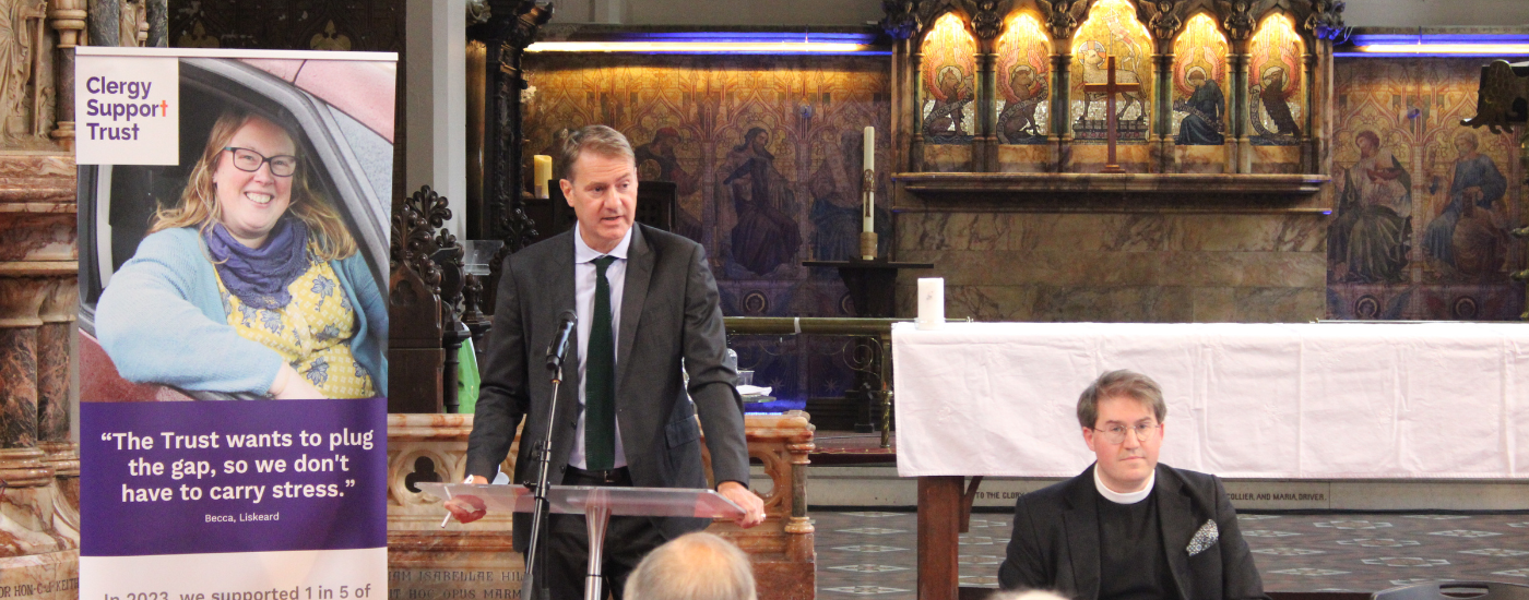 One man, dressed in a suit and delivering a speech behind a podium, and another, sat behind a table.