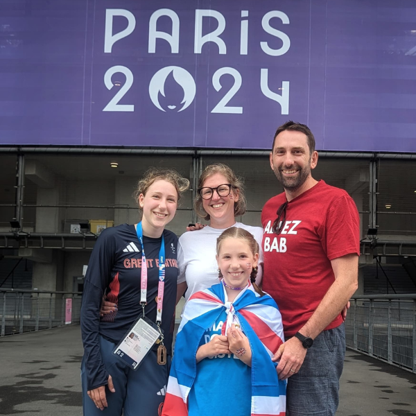 Sarah, her husband and two daughters, standing in front of a Paris 2024 sign.