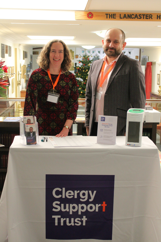 A man and a woman smiling, stood behind a table cloth with the Clergy Support Trust logo.