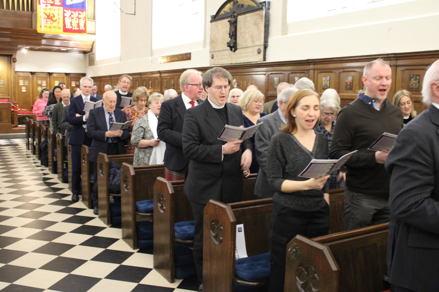 A chequered floor of white and black tiles. The chapel's organ can be seen in the background. Guests are singing from the Order of Service.