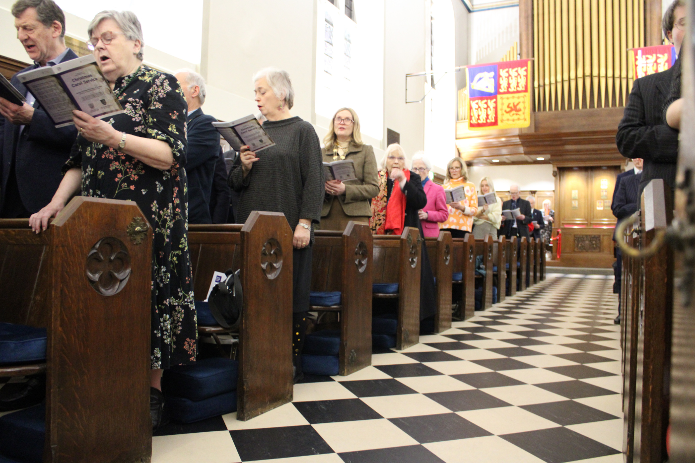 A chequered floor of white and black tiles. The chapel's organ can be seen in the background. Guests are singing from the Order of Service.