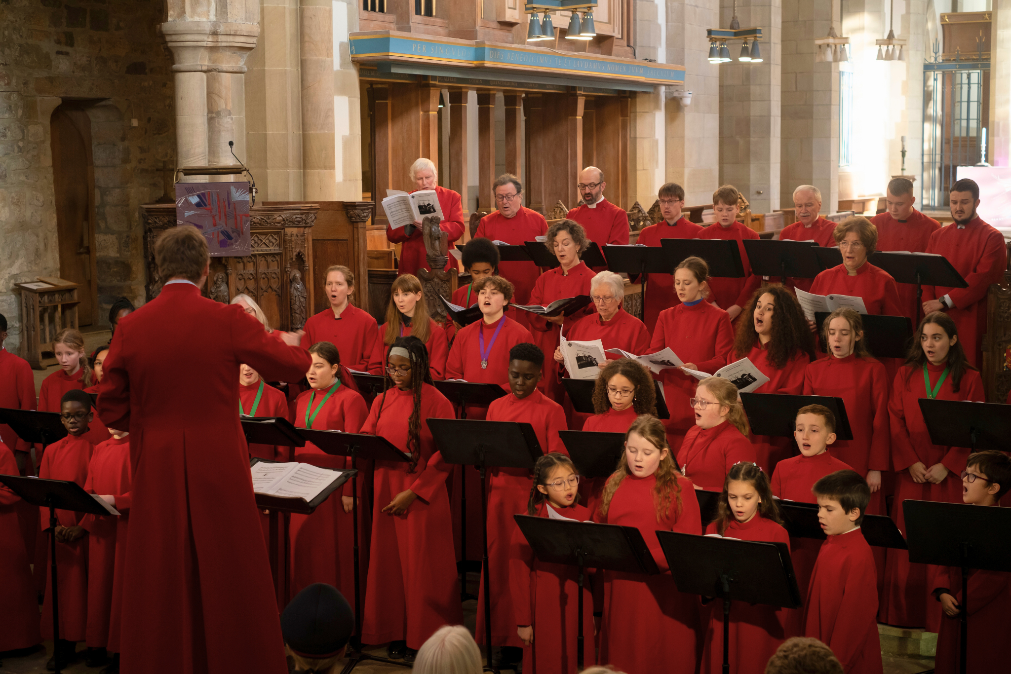 Several lines of young choristers standing and singing, dressed in red robes, and led by their Director of Music.