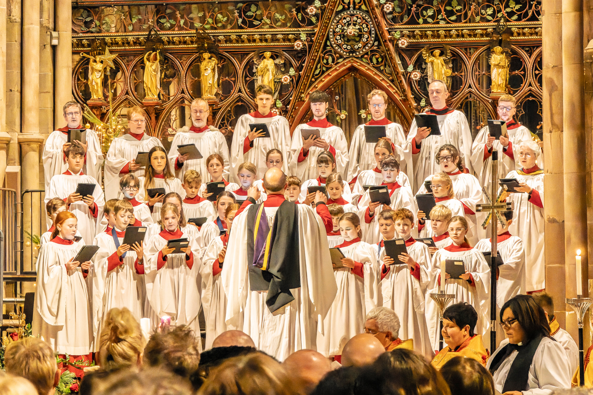 Several lines of young choristers standing and singing, dressed in white robes, and led by their Director of Music.