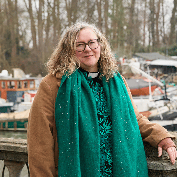 A woman, standing in front of a harbour, wearing a clerical collar, glasses and a green dress. Moored boats can be seen in the background.