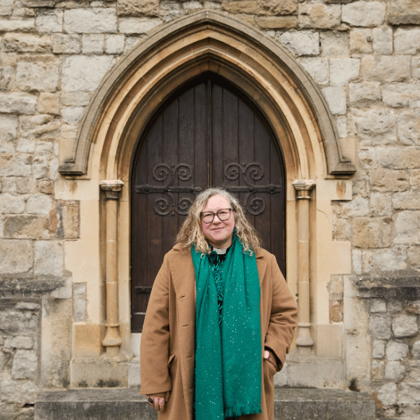 A woman, standing in front of a church's wooden door, wearing a clerical collar, glasses and a brown coat.