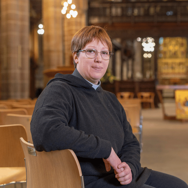 Kathryn is wearing a black top and a white clerical collar. She is sat on a wooden chair.