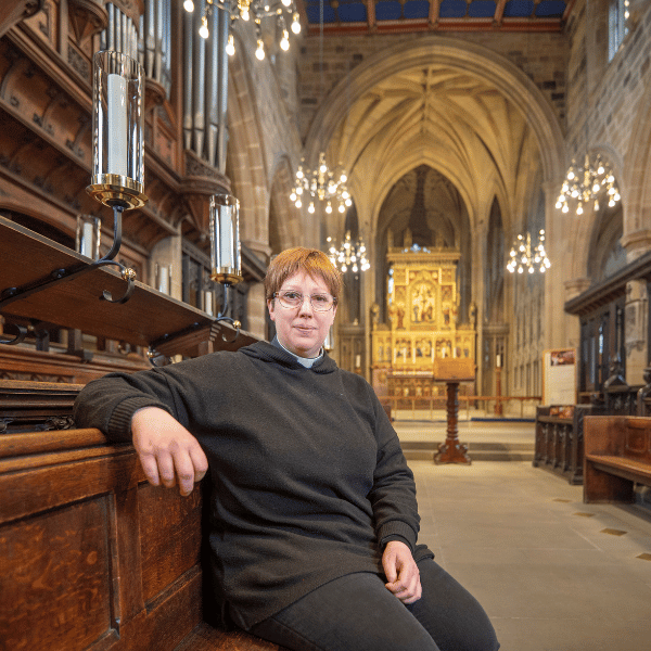 Kathryn is wearing a black top and a white clerical collar. She is sat on a wooden pew, with candles hung from the ceiling behind her.