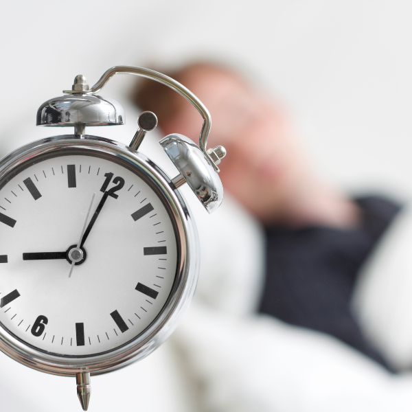 A silver bed-side clock, and a blurred image of a man sleeping in a bed in the background.