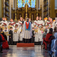 A choirmaster stands in front of the choir, conducting them, to a seated audience,