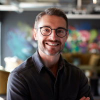 A man, wearing a dark t-shirt and glasses, looking at the camera and smiling.