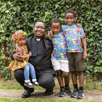 Josiah, standing in front of a hedge, smiling with his daughter and two sons.