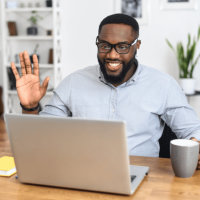 A man wearing glasses and a light blue shirt, sat at his desk and waving to someone who he is calling on his laptop.