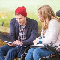 A man and a woman in a park, reading a page inside a folder. The man is sat on a bench, while the women is sat on a motorised wheelchair.