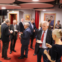 A group of people, dressed formally, standing and talking with hot drinks.